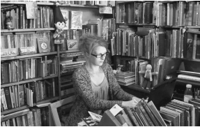 photo of Jen sitting at a desk in a bookshop, surrounded by bookshelves. She is typing at a computer.